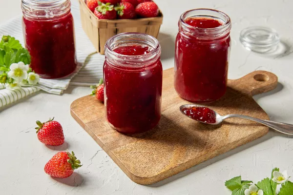 Three jars of strawberry jam, two on a wood cutting board and one on a tea towel, shown with a spoon of jam, fresh strawberries, and strawberry flowers.