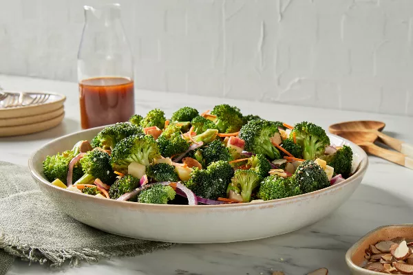 A serving bowl of broccoli salad with gochujang dressing on a kitchen counter shown with a pitcher of dressing, plates, and wooden serving spoons.