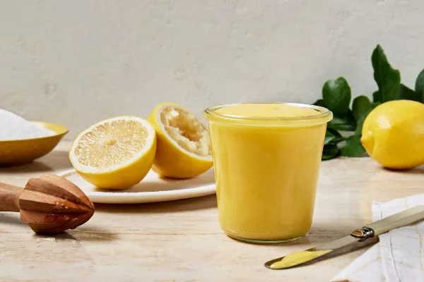  A glass jar full of lemon curd shown with a bisected lemon with one half juiced, a lemon juicer, and a bowl of granulated sugar.