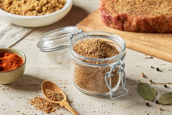 A lidded glass jar full of sweet Montreal steak seasoning shown with a raw, seasoned steak on a cutting board, a bowl of brown sugar, and a bowl of spices.