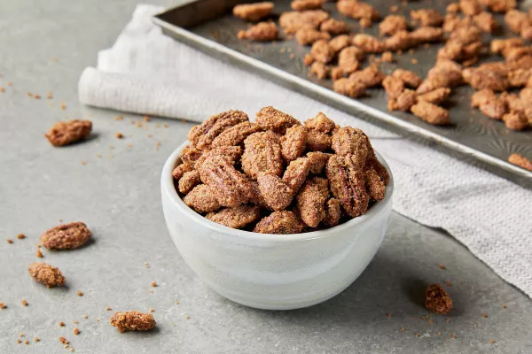 A white bowl overflowing with candied pecans and almonds, shown on a kitchen counter with a baking sheet of prepared nuts.