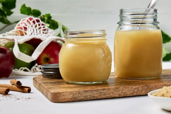 Two glass jars of homemade applesauce on a wooden cutting board on a white surface, shown with a mesh bag of apples, cinnamon sticks, and a bowl of Golden Yellow Sugar.
