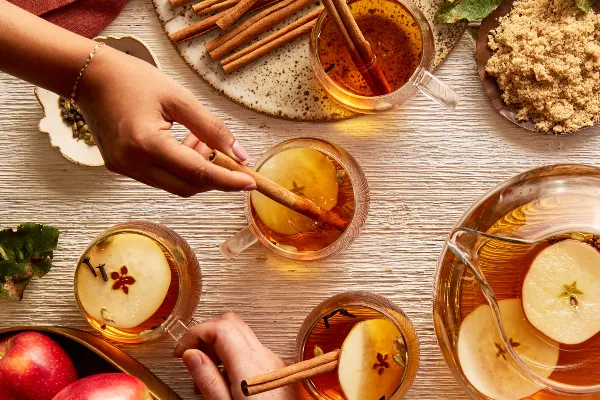 Four glass mugs of hot apple cider garnished with apple slices, cloves, and cinnamon sticks, shown on a table with a pitcher of cider, a bowl of apples, and a bowl of golden yellow sugar, and hands reaching for the mugs.