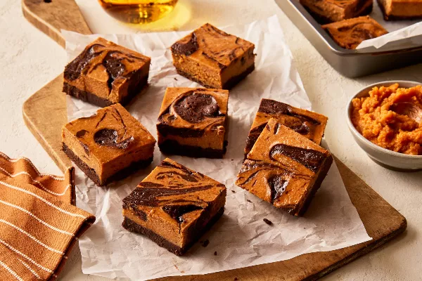 Seven pumpkin cheesecake brownies on a wooden cutting board lined with parchment, shown with a baking pan with brownies and a bowl of pumpkin puree.