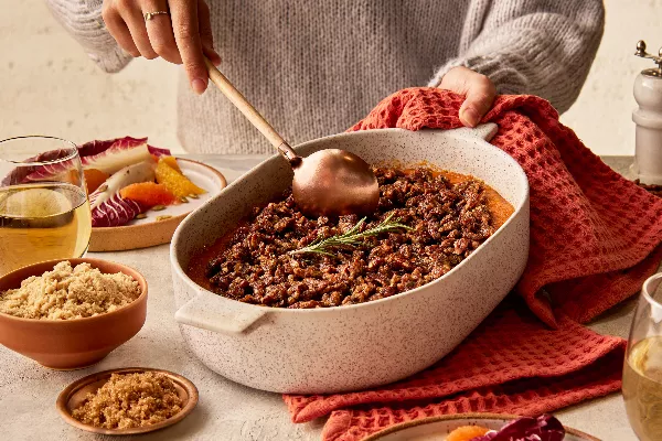 A woman wearing a sweater serving sweet potato casserole garnished with a rosemary sprig from a baking dish, shown with a radicchio and citrus salad, glasses of wine, and bowls of golden yellow and demerara sugar.