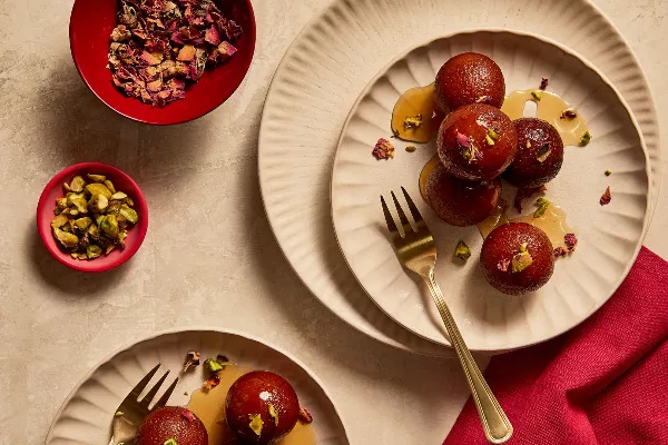 Balls of fried dough (Gulab Jamuns) with golden forks and small bowls filled with petals and pistachio