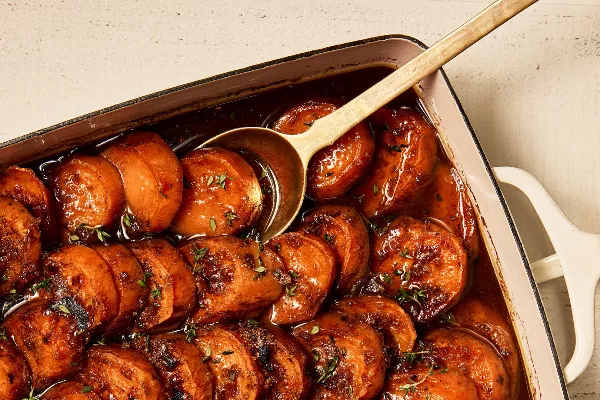 Sliced candied yams shown in a baking dish with a serving spoon, garnished with herbs and showing visible caramelization.