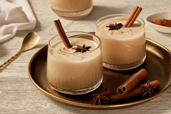 Two tumbler glasses of eggless eggnog garnished with anise, cinnamon, and cinnamon sticks, shown on a gold tray with anise and cinnamon sticks, and a gold stirring spoon beside