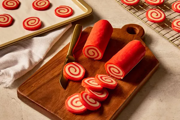  Two rolls of red and white peppermint pinwheel cookie dough on a wooden cutting board with several slices cut, with more slices on a baking sheet and baked cookies cooling on a wire rack.