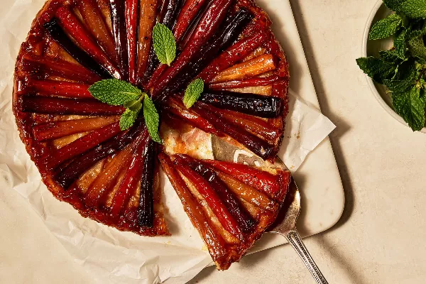 A carrot and parsnip tarte tatin resting on wax paper on a cutting board, shown with one slice partially removed with a pie lifter, garnished with fresh mint leaves.