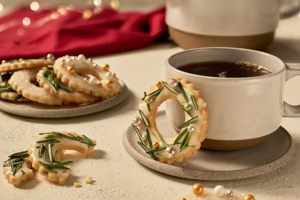 Glazed sugar cookies with icing and decorated like wreaths with rosemary, one on a saucer with a cup of coffee, one broken on a table, and several on a plate, shown in a festive holiday setting.