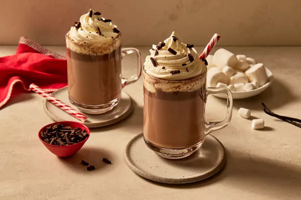 Two glass mugs of vanilla hot chocolate topped with whipped cream and garnished with shaved chocolate and candy canes, resting on saucers and shown with marshmallows, shaved chocolate, and vanilla beans. 