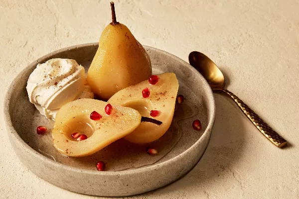 Three halves of white wine poached pears shown in a shallow dish, served with a dollop of whipped cream and sprinkled with cinnamon and pomegranate arils, and shown with a gold spoon.
