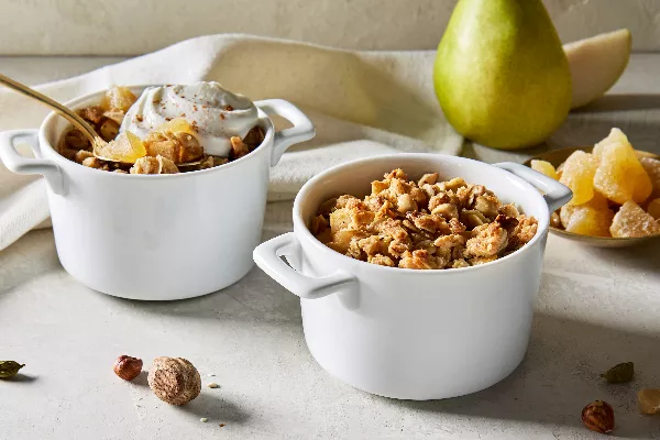 Two white single-serving bowls of pear ginger crumble, one with a dollop of whipped cream and a gold spoon, shown with a whole pear and a small dish of ginger pieces.