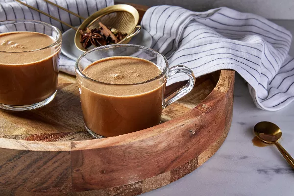 Two glass mugs of masala chai on a round wooden serving tray, shown with a gold strainer with whole spices in it, a dishcloth, and a gold spoon.