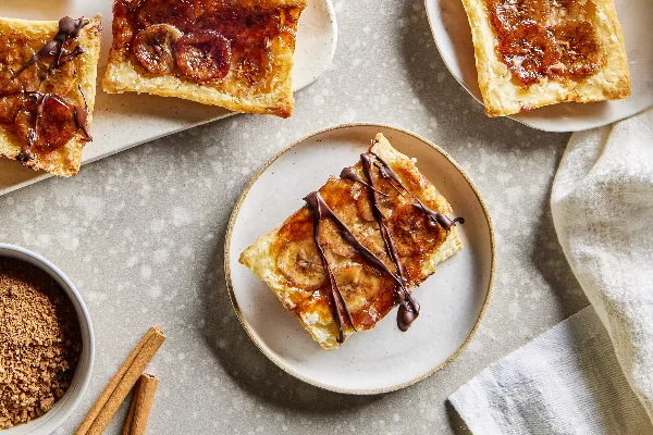 Four upside-down banana tarts, two on plates and two on a tray, shown with a bowl of brown sugar and cinnamon sticks.