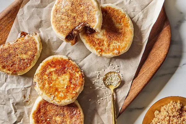 Five Korean pancakes or hotteok on a wood tray, two cut open with sesame syrup leaking out, shown with scattered sesame seeds and a bowl of Demerara Style Sugar.