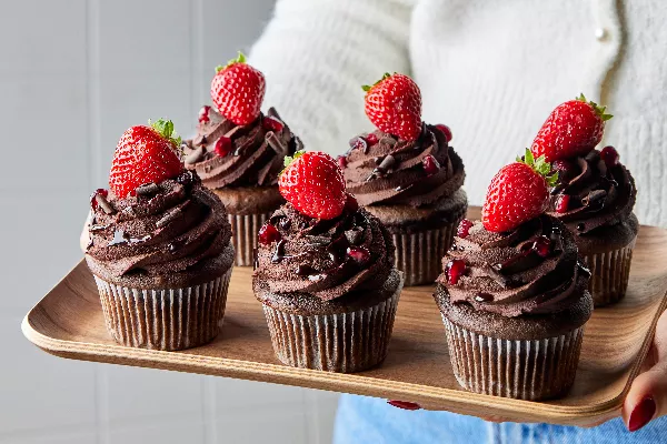 Six red wine chocolate cupcakes topped with ganache, shaved chocolate, pomegranate, and strawberries on a wooden tray held by a woman in a white sweater and jeans
