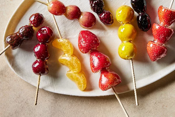 A platter of skewered fruit and berries coated in clear sugar syrup, or tanghulu, including mandarin segments, cherries, grapes, gooseberries, strawberries, kumquats, and blackberries