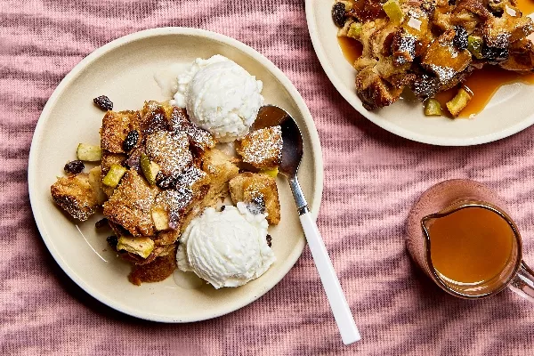 A serving of apple bread pudding on a plate with visible raisins and apple pieces, served with vanilla ice cream, shown with a second plate of bread pudding with butterscotch sauce and a glass pitcher of sauce.