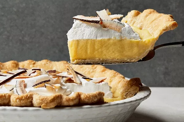  A slice of coconut cream pie shown from the side on a pie lifter, freshly cut from a full pie in a decorative white baking dish, topped with whipped cream and shaved coconut.