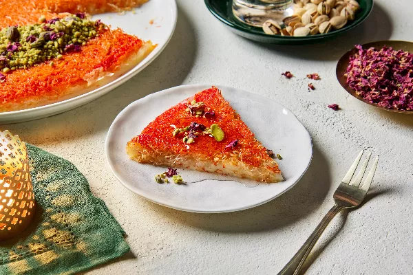 A slice of knafeh on a plate, garnished with pistachios and edible flower petals, shown with a full knafeh, dishes of pistachios in the shell and flower petals and a lit candle. 