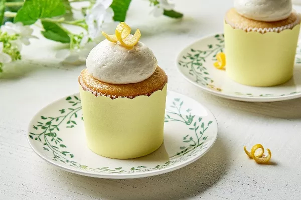 Lemon cupcakes with a fluffy vanilla frosting, adorned with a twist of lemon zest, presented on a vintage floral-patterned plate set against a white wooden surface and a backdrop of soft greenery and white blooms for a fresh, springtime feel.