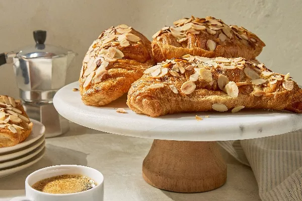 Four air fryer almond croissants topped with sliced almonds on a white cake stand, with an espresso cup and stovetop coffee maker in the background.