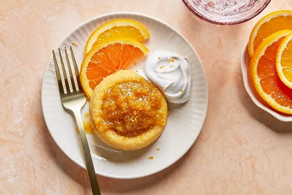 Overhead shot of a single sponge cake topped with glistening citrus marmalade and zest, served on a white ridged plate with fresh orange slices and a dollop of whipped cream, accompanied by a fork, against a soft peach-toned backdrop.