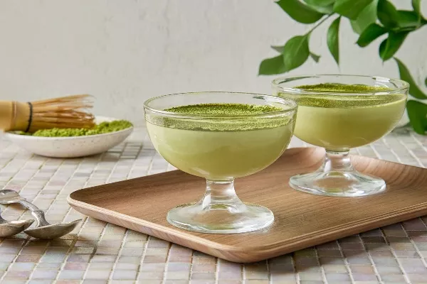  Two glass cups of white chocolate matcha mousse garnished with green matcha powder on a wooden tray, with a traditional matcha whisk and bowl of matcha powder in the background, set on a tiled table with natural light.