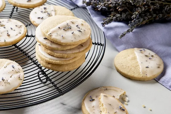 A batch of glazed lemon-lavender shortbread cookies sprinkled with dried lavender, shown on a circular wire cooling rack on a marble counter, with dried lavender stalks and a lavender-coloured cloth.