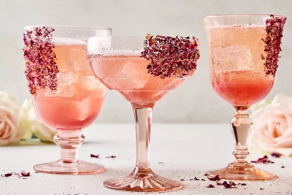Three unique stemmed glasses of lychee-rose spritzer on ice garnished with dried rose petals on the glasses, shown in a neutral setting with fresh roses in the background and dried rose petals scattered around.