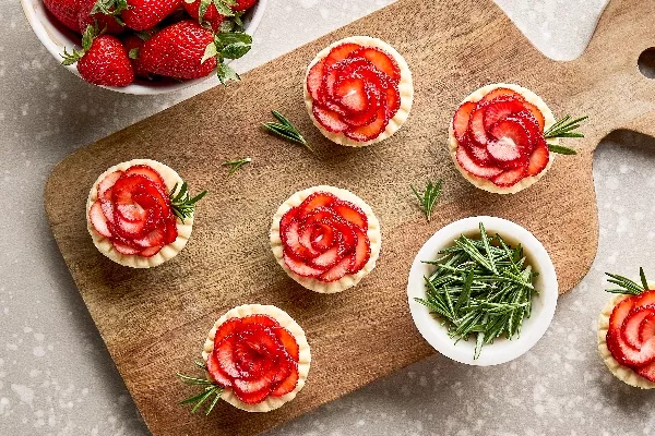 Five strawberry rosemary tartlets on a wooden cutting board, shown with a bowl of rosemary sprigs, a bowl of fresh strawberries, and a sixth tart on the counter.
