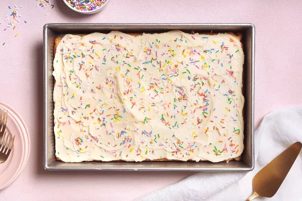 A rectangular frosted cake covered in colorful sprinkles sits in a metal baking pan on a light pink surface, with a small bowl of more sprinkles, plates and forks, and a gold cake server nearby.