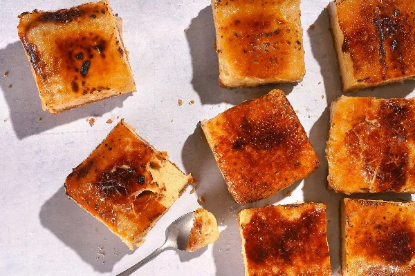 An overhead shot of several small, square slices of caramelized cheesecake with a burnt sugar topping, scattered on a white surface with a spoon taking a bite out of one piece.