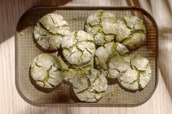 Eight pale green matcha crinkle cookies, coated in powdered sugar with characteristic cracks, arranged on a rectangular metal baking sheet.