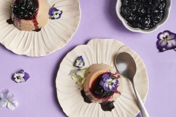  Top-down view of two servings of Earl Grey panna cotta topped with blueberry compote and decorated with purple and white edible flowers, served on scalloped cream plates against a lavender background with scattered blossoms and a spoon.