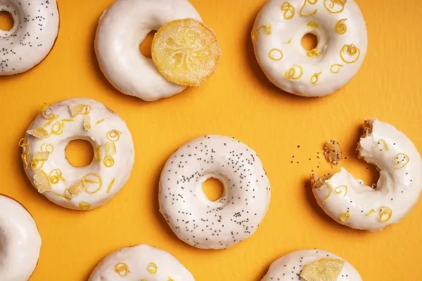 Glazed lemon poppyseed doughnuts arranged on a vibrant yellow background, decorated with lemon zest, candied lemon slices, and poppy seeds; one doughnut has a bite taken out.