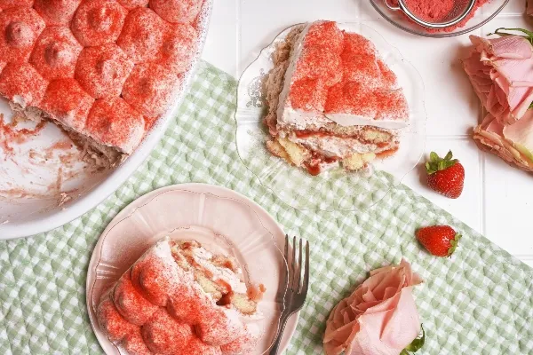 A white baking dish of strawberries and cream tiramisu on a green gingham cloth on a white tiled surface with two slices cut out and served on pink and clear glass plates, surrounded by fresh strawberries and pink roses.