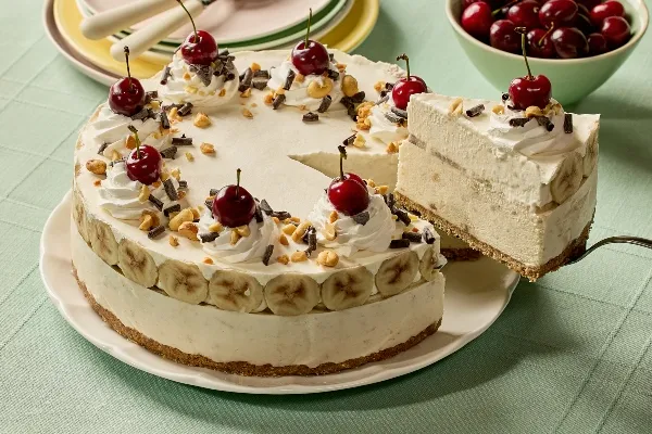 A creamy banana split ice cream pie topped with swirls of whipped cream, cherries, chopped nuts, and chocolate shavings sits on a white plate. A slice is being lifted to reveal layers of banana slices and frozen filling over a graham cracker crust. In the background, there’s a stack of pastel plates with forks and a bowl of fresh cherries on a light green tablecloth.