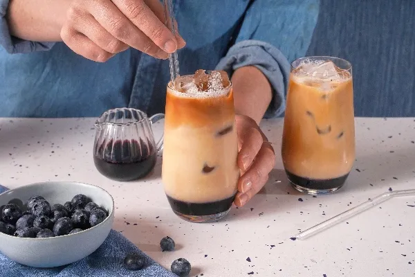 Person in a denim shirt stirring a glass of iced blueberry coffee with a glass straw. The drink has distinct layers of blueberry syrup, milk, and coffee. A second identical drink sits nearby on a speckled countertop. A glass pitcher of blueberry syrup, a bowl of fresh blueberries, and scattered berries complete the scene.