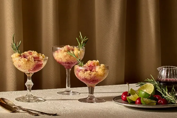 Three coupe glasses filled with a layered frozen dessert of golden brown and ruby red slush, garnished with fresh rosemary sprigs, are set on a light textured surface in front of a warm brown curtain. Beside them sits a plate with fresh limes, lime wedges, cherries, and rosemary, as well as a small glass pitcher of dark cherry syrup