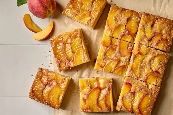 Top-down view of golden-brown peach upside-down cake squares arranged on a sheet of parchment paper. The squares are topped with glistening caramelized peach slices. A whole fresh peach and a few peach wedges are placed decoratively beside the cake pieces.