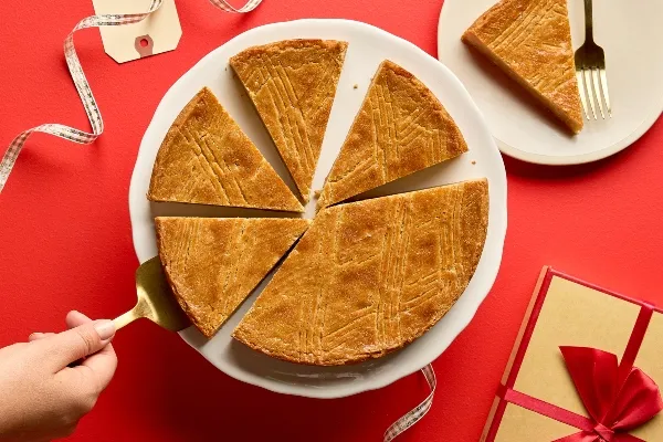 Sliced Brown Sugar Dutch Butter Cake on a white cake stand against a red background. One slice is being lifted with a gold cake server. A single slice sits on a small white plate with a gold fork, and a wrapped gift box and ribbon are nearby.