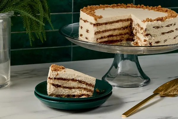 A layered gingersnap icebox cake on a glass cake stand, with visible layers of spiced cookie crumbs and creamy filling. A slice is served on a dark green plate in the foreground, alongside a gold cake server on a marble countertop.