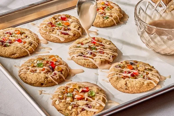 Freshly baked fruitcake cookies on a parchment-lined baking sheet, topped with colourful candied fruit and being drizzled with a glossy glaze from a spoon. A bowl of glaze sits nearby.