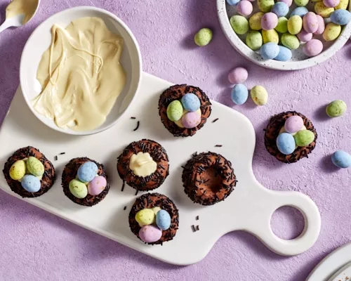 Seven chocolate bird nest cookies topped with white chocolate and candy-coated chocolate eggs on a cutting board