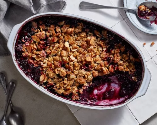 Serving dish full of blueberry crisp with a small amount scooped out on a tile counter