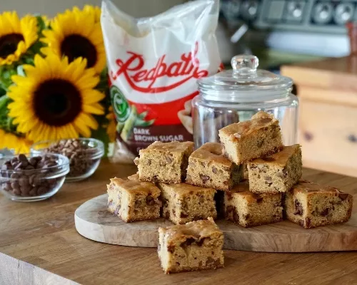 Brown Butter Blondies with chocolate chips on a butcher block counter with sunflowers and a bag of Redpath Dark Brown Sugar