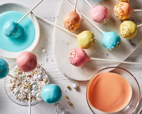 Pastel-coloured cake pops with sprinkles on a platter, some standing in a glass of sprinkles, and one being dipped in blue icing